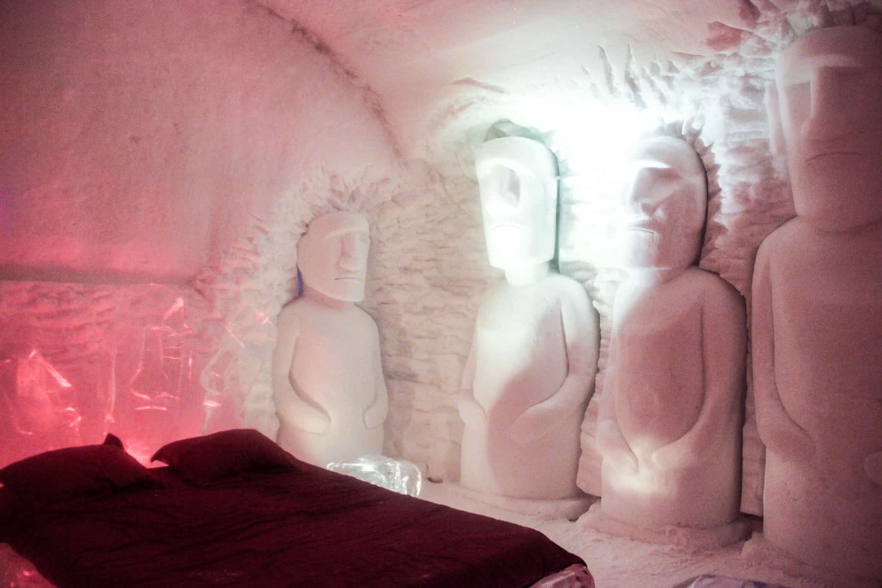 A bed with a red duvet and pillows and four figures carved from ice at Quebec's ice hotel, Hôtel de Glace.