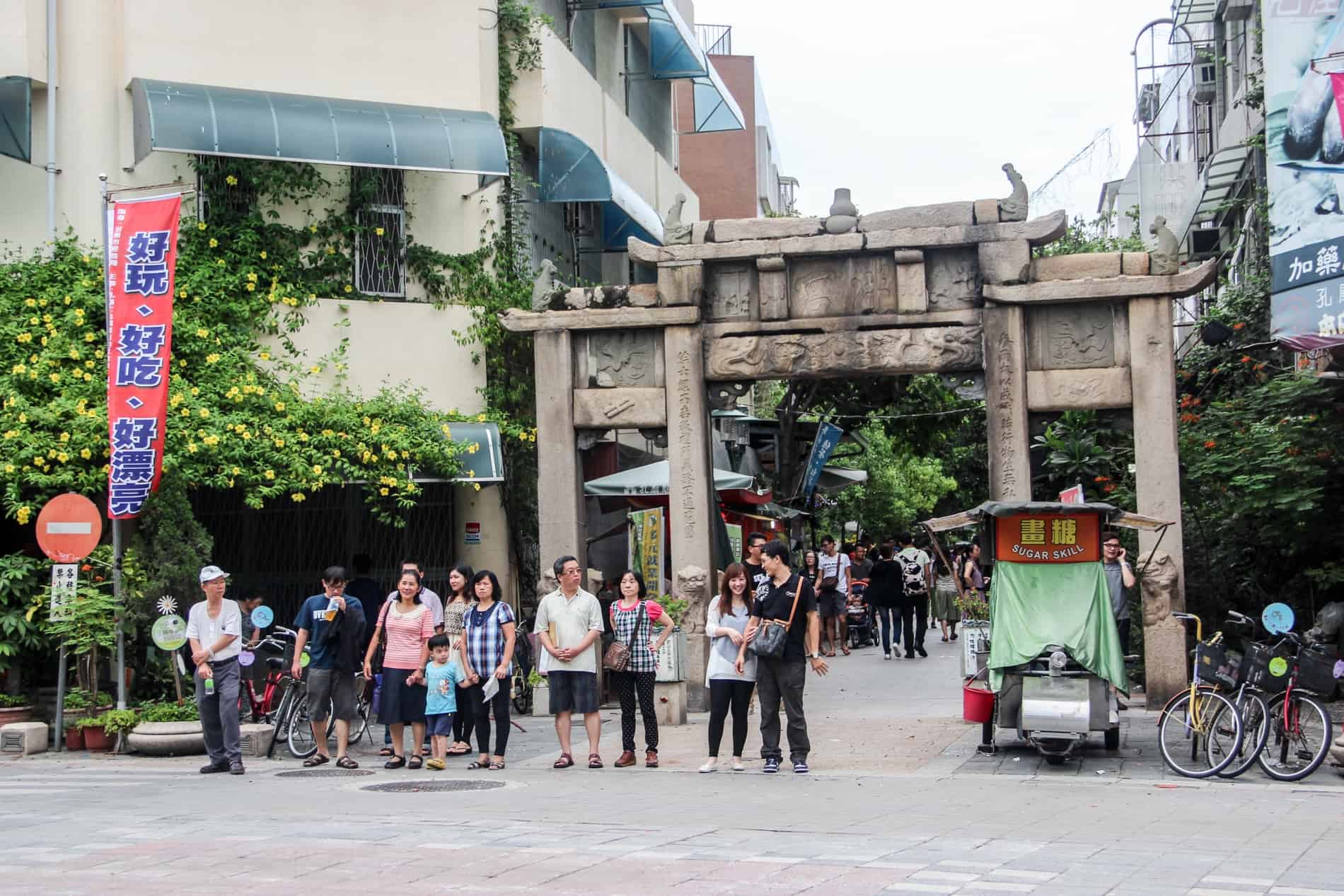 People standing in a line on a street in front of an old Chinese stone gate. 
