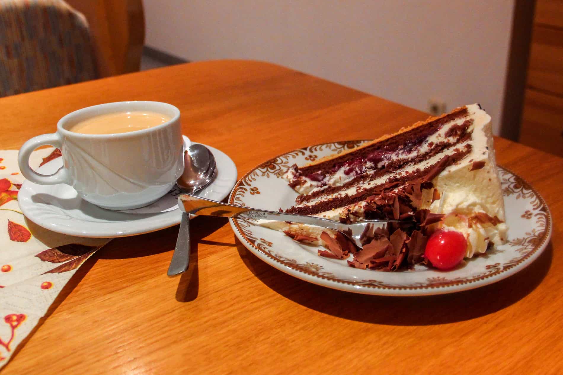 A cup of coffee and a slice of Black Forest Gateau - a dark, layered sponge cake covered in cream, and topped with a cherry. 