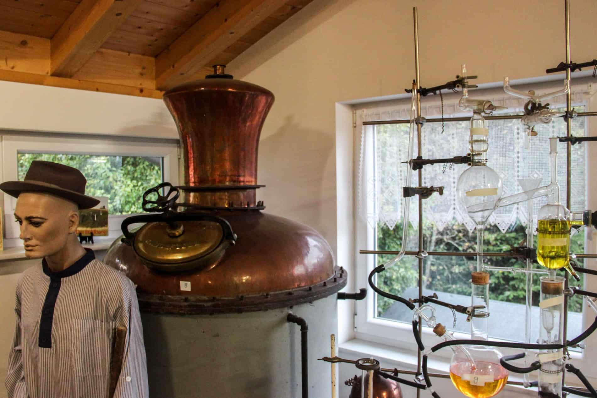 A mannequin of a distillery worker in front of of a copper fermentation vats and a stack of glass tubes. 