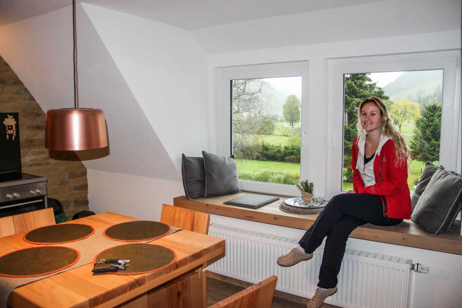 A blonde woman sits on a wooden window seat of a kitchen-dining room in a house with forest views. 