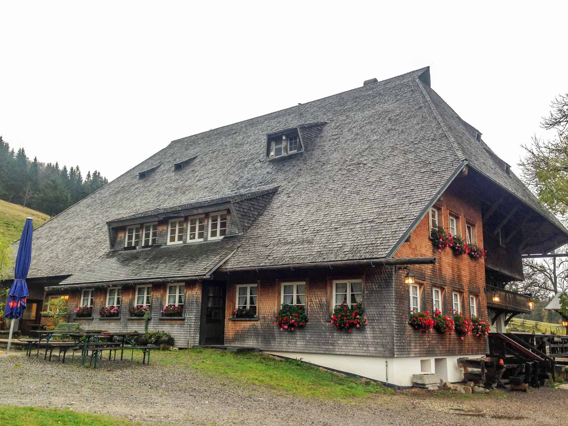 A large German alpine house in the Black Forest with blackened orange tiles, flower pot window balconies, and a grey tiled roof. 