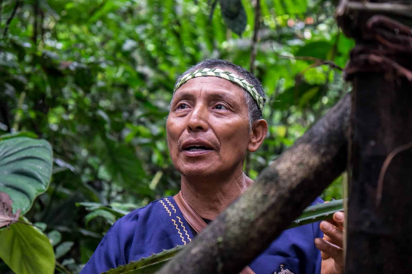 Local man, Delfin wearing a headband weaved from leaves, holds a long plant while standing in the jungle of the Ecuador Amazon Rainforest.