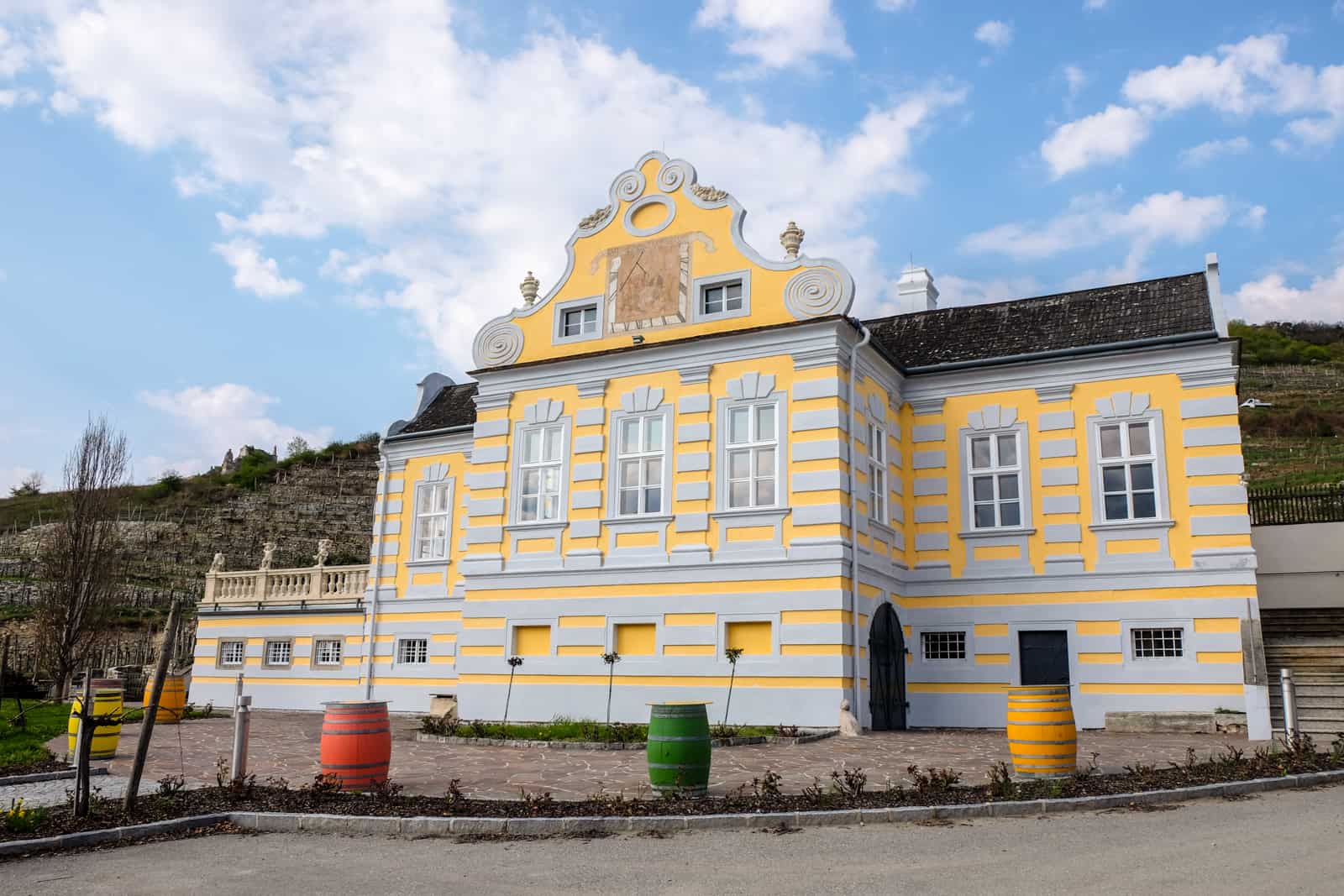 A yellow and grey classic looking building in a wine valley in Austria with yellow, red and green barrels outside 