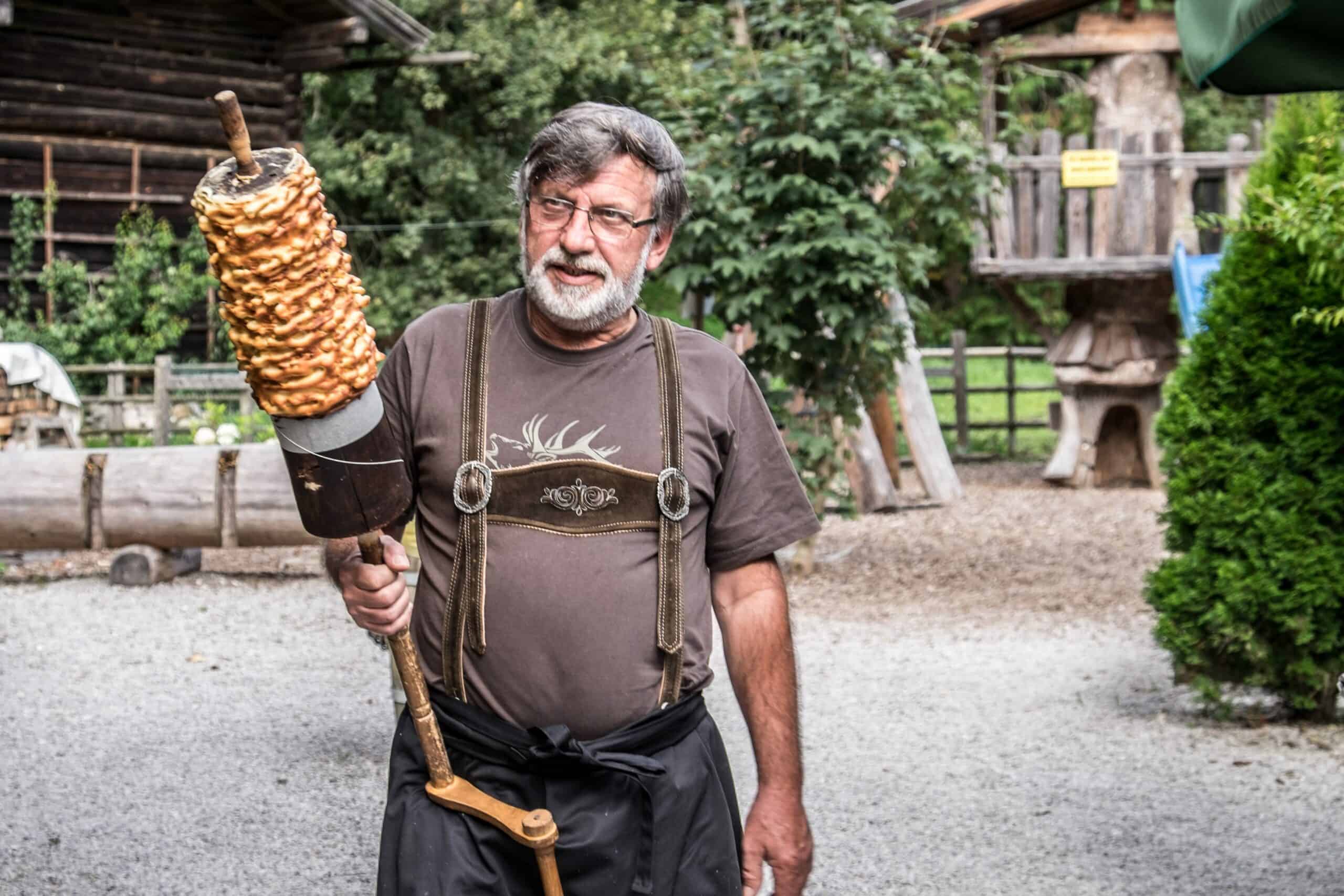 A man dressed in a Lederhosen top carries a pole covered with a large cylindrical cake. 