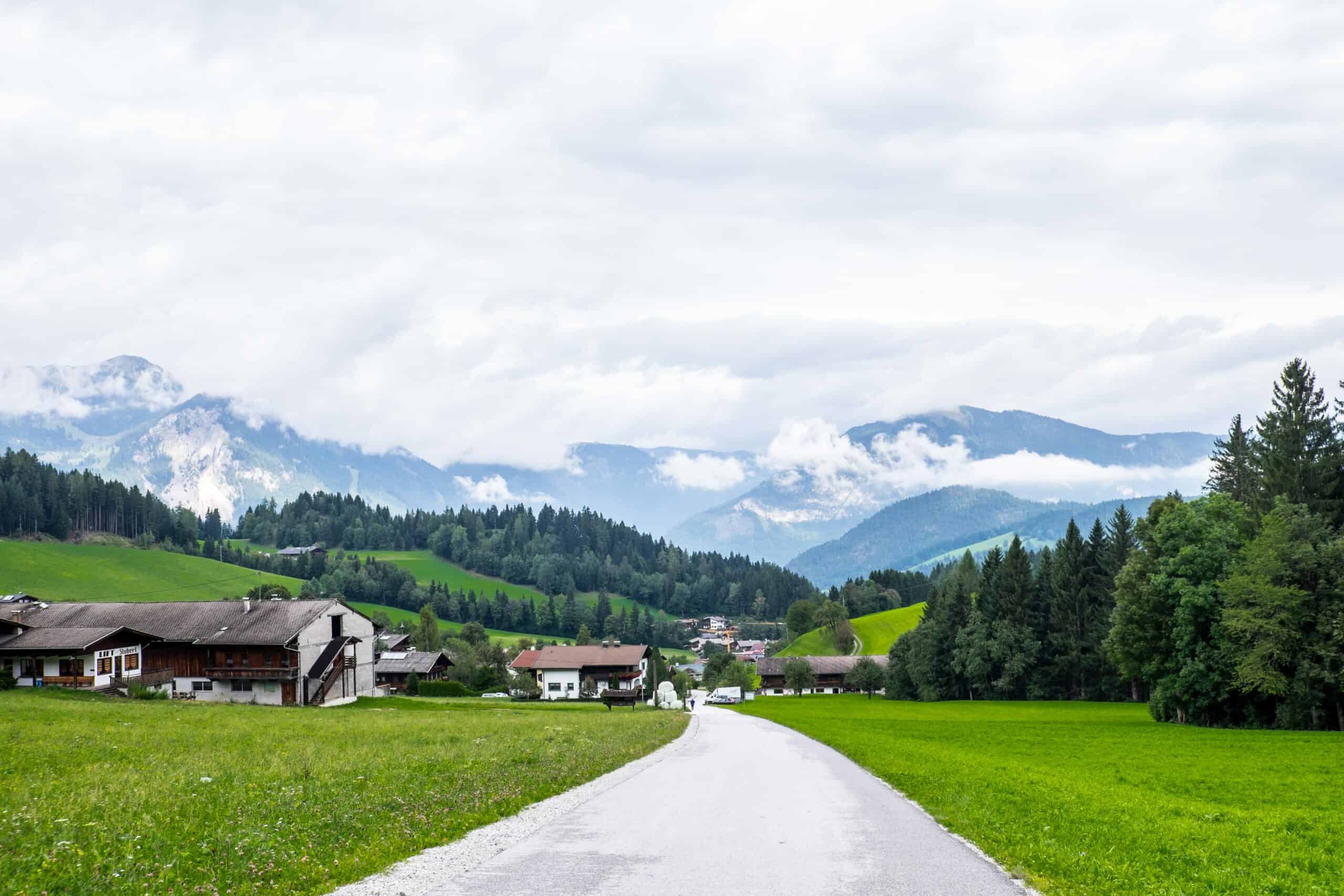 View down a grey path towards an alpine village in front of a low-slope forest and mountains. 