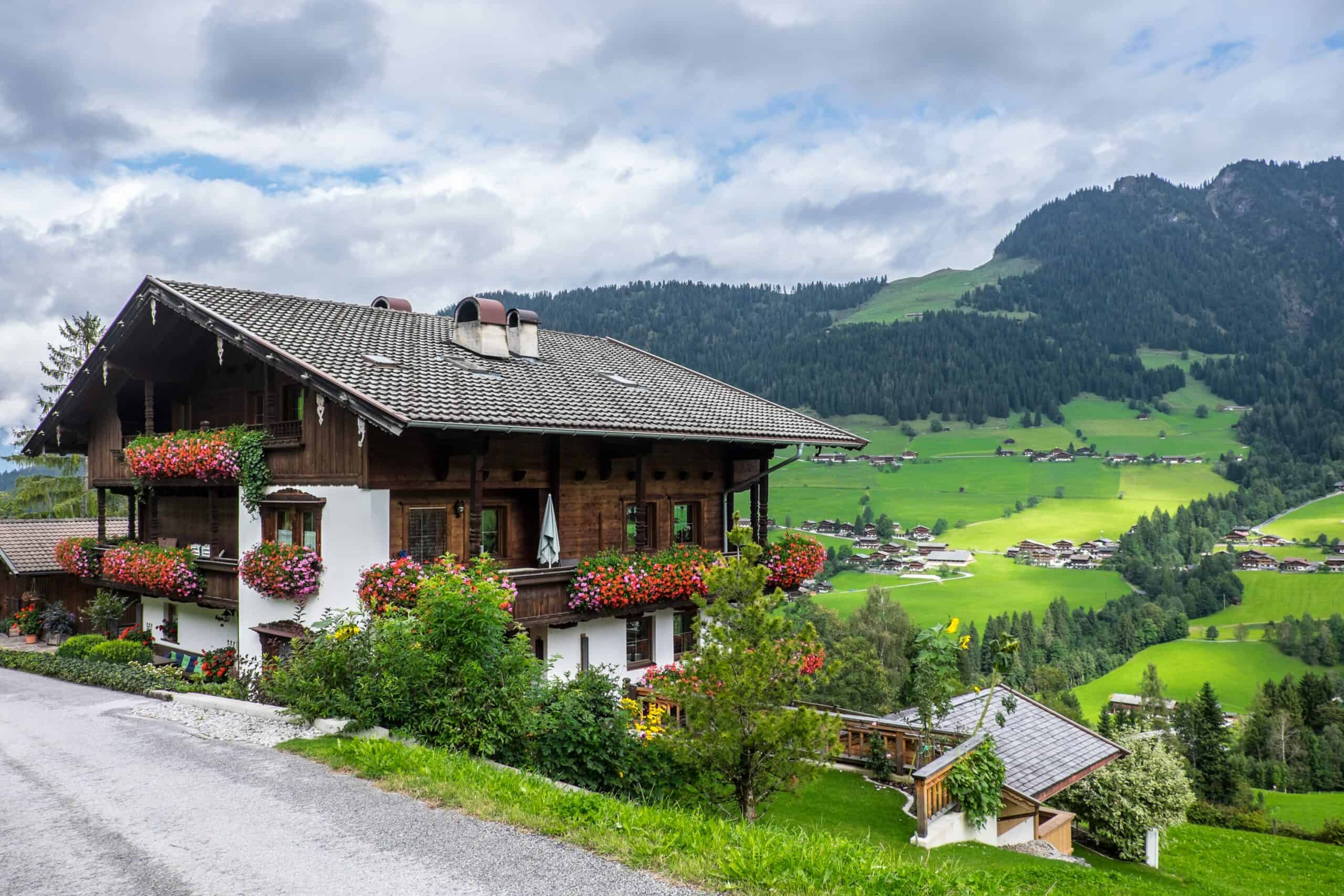 A wooden Austrian alpine house with flower-filled balconies perched above a valley dotted with houses. 