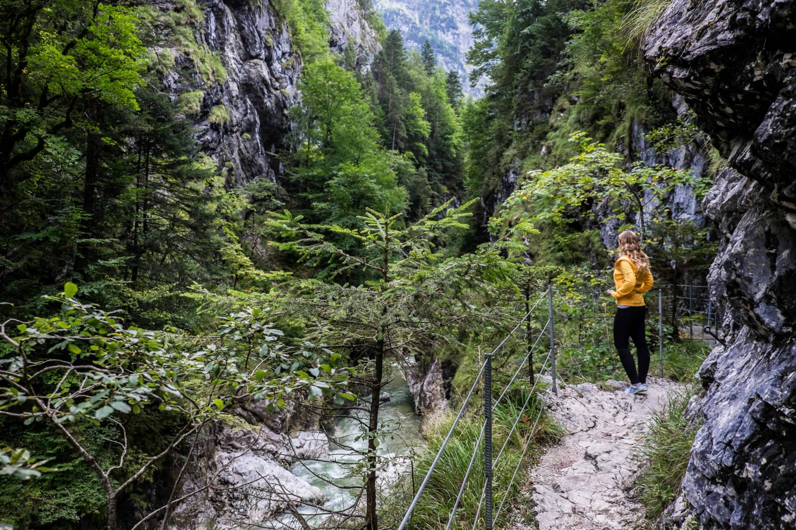 A woman in a yellow jacket stands on a rock pathway in a deep, foliage filled gorge looking out over a river.