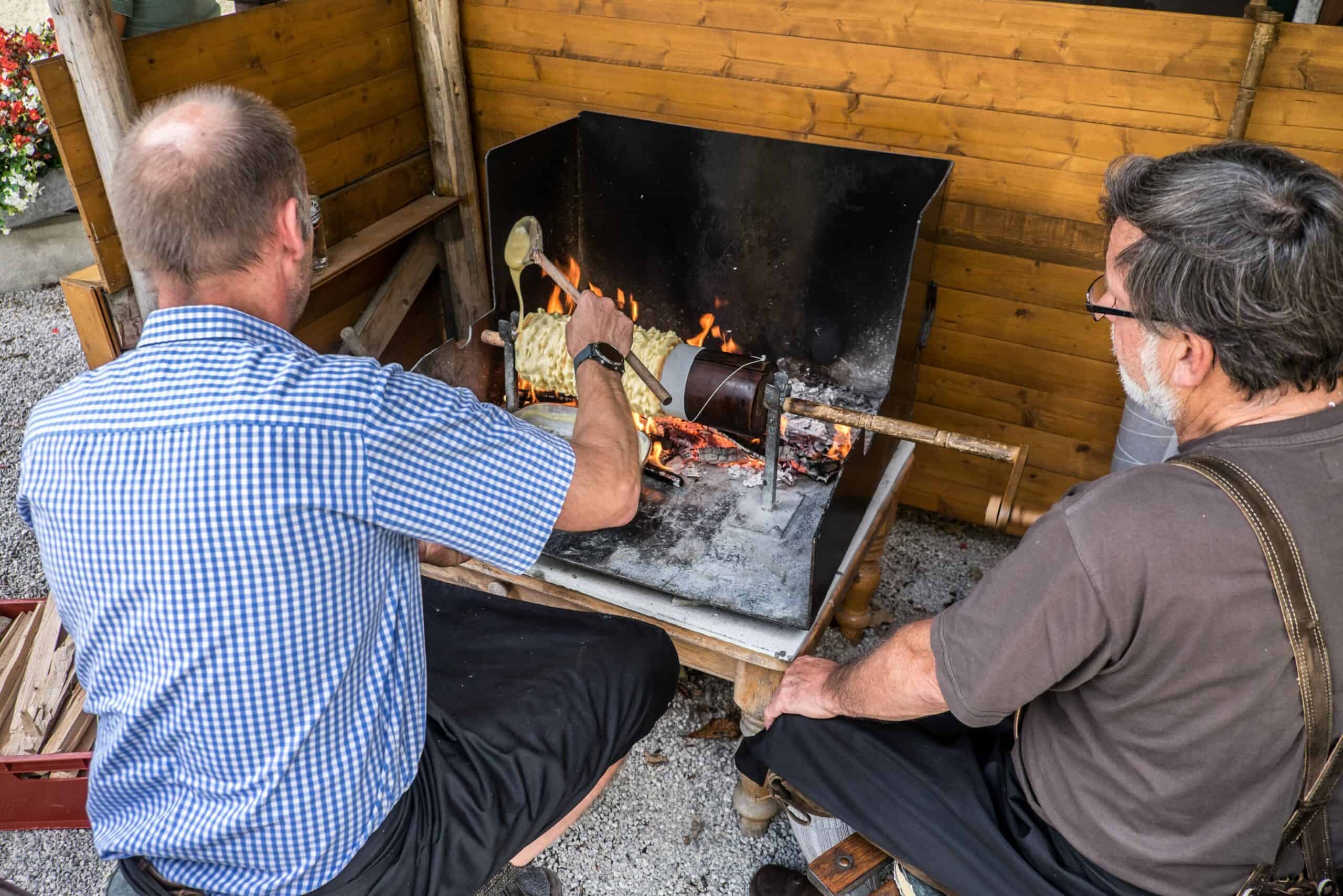 Two men sit in front of a small grill; the man in checkered blue shirt pours a mixture over a cylindrical spinner. 