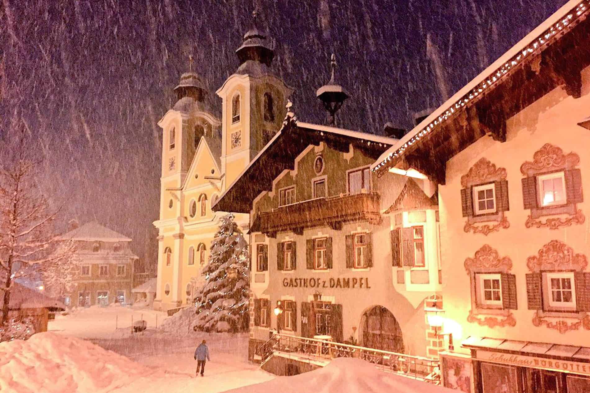 Snow falls over the painted facades of traditional Austrian mountain village huts and the church in St. Johann in Tirol. 