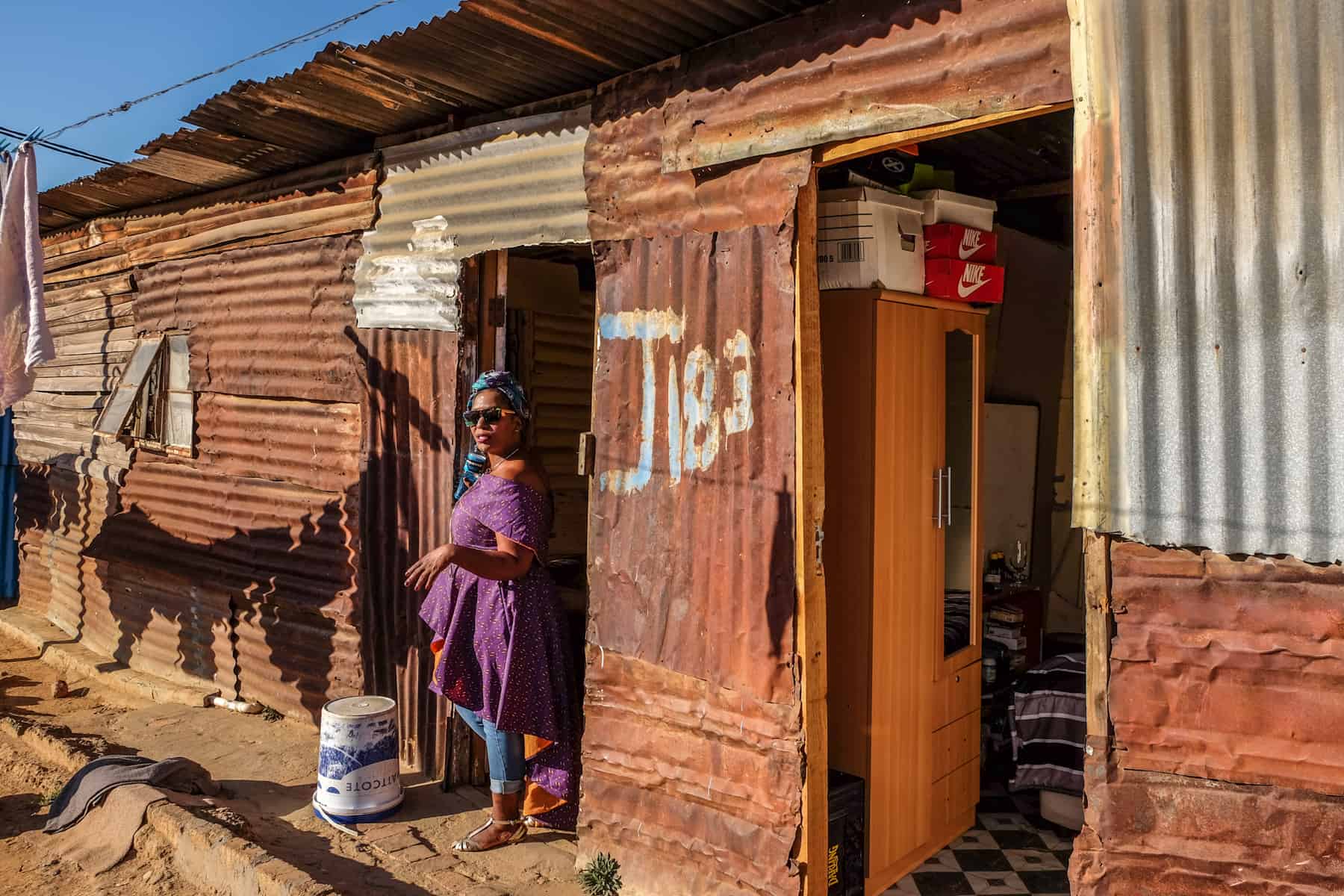 A local woman in a purple dress stands in the doorway of a clay coloured one-floor housing unit in a township. She is guiding a tour group through her home in Kayamandi 