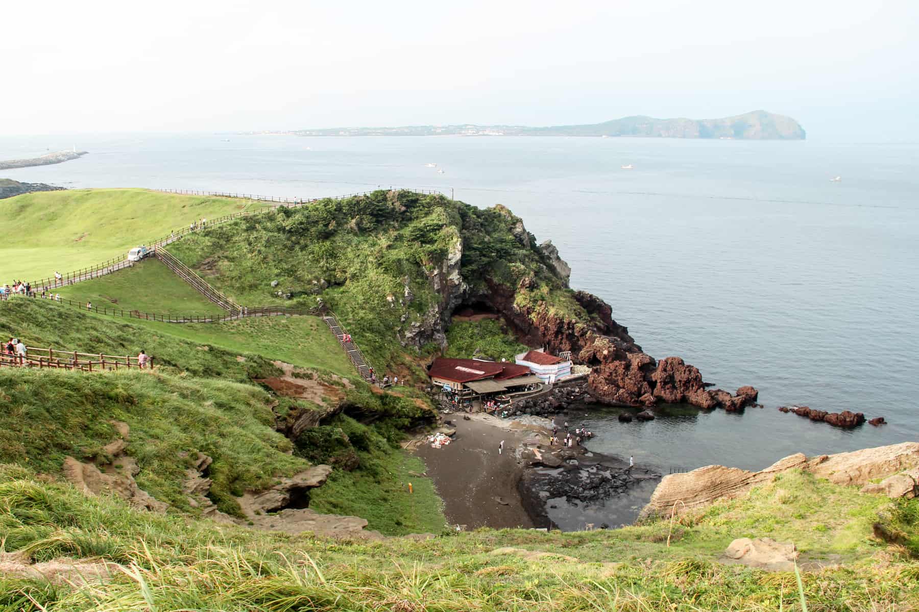 People walking on narrow paths on a green mound of Jeju Island's Sunrise Peak, looking down onto red-roofed houses, coastal rocks and the vast ocean.