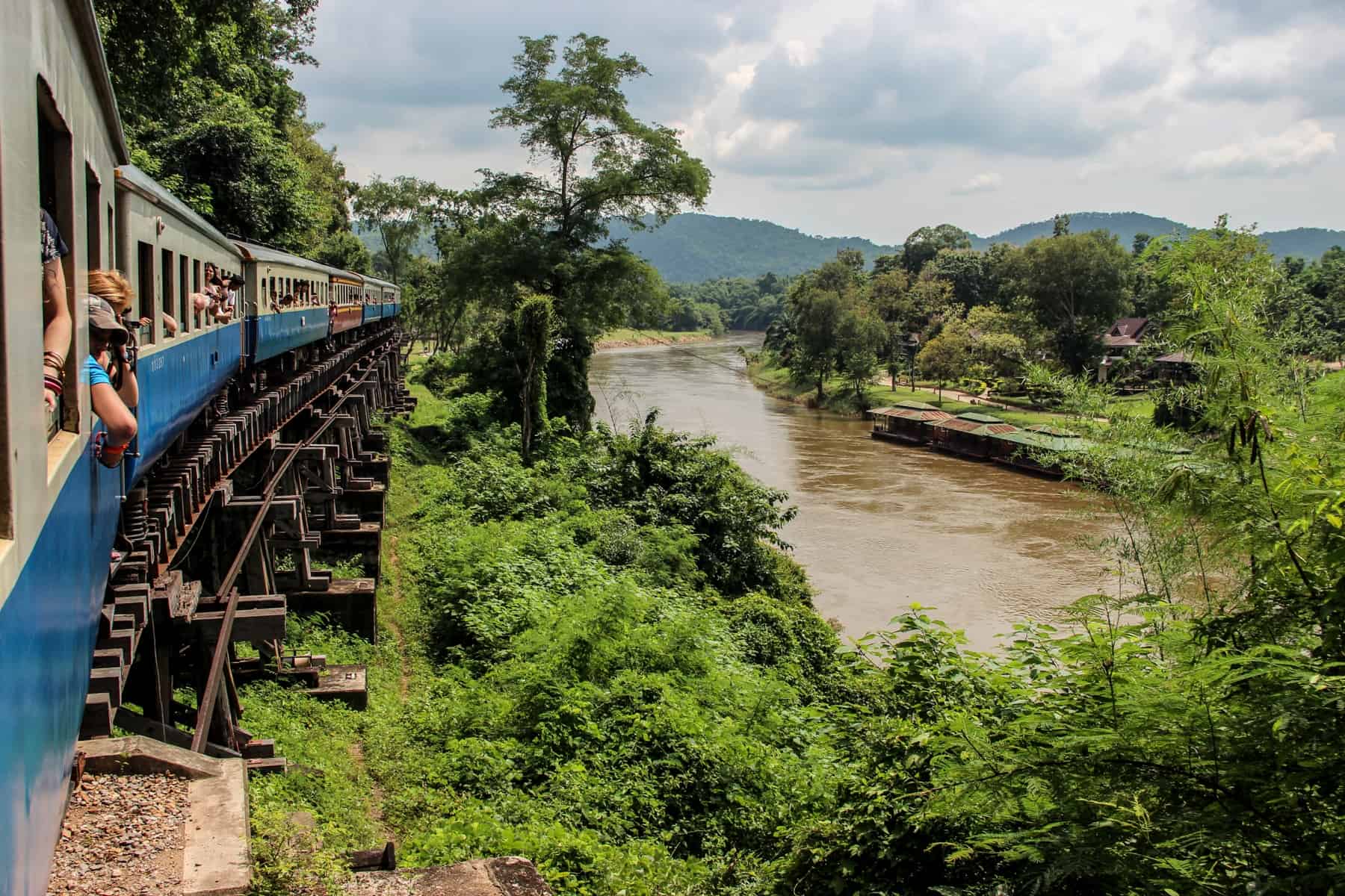 A white and blue train passes a river in a green jungle valley in Kanchanaburi, Thailand