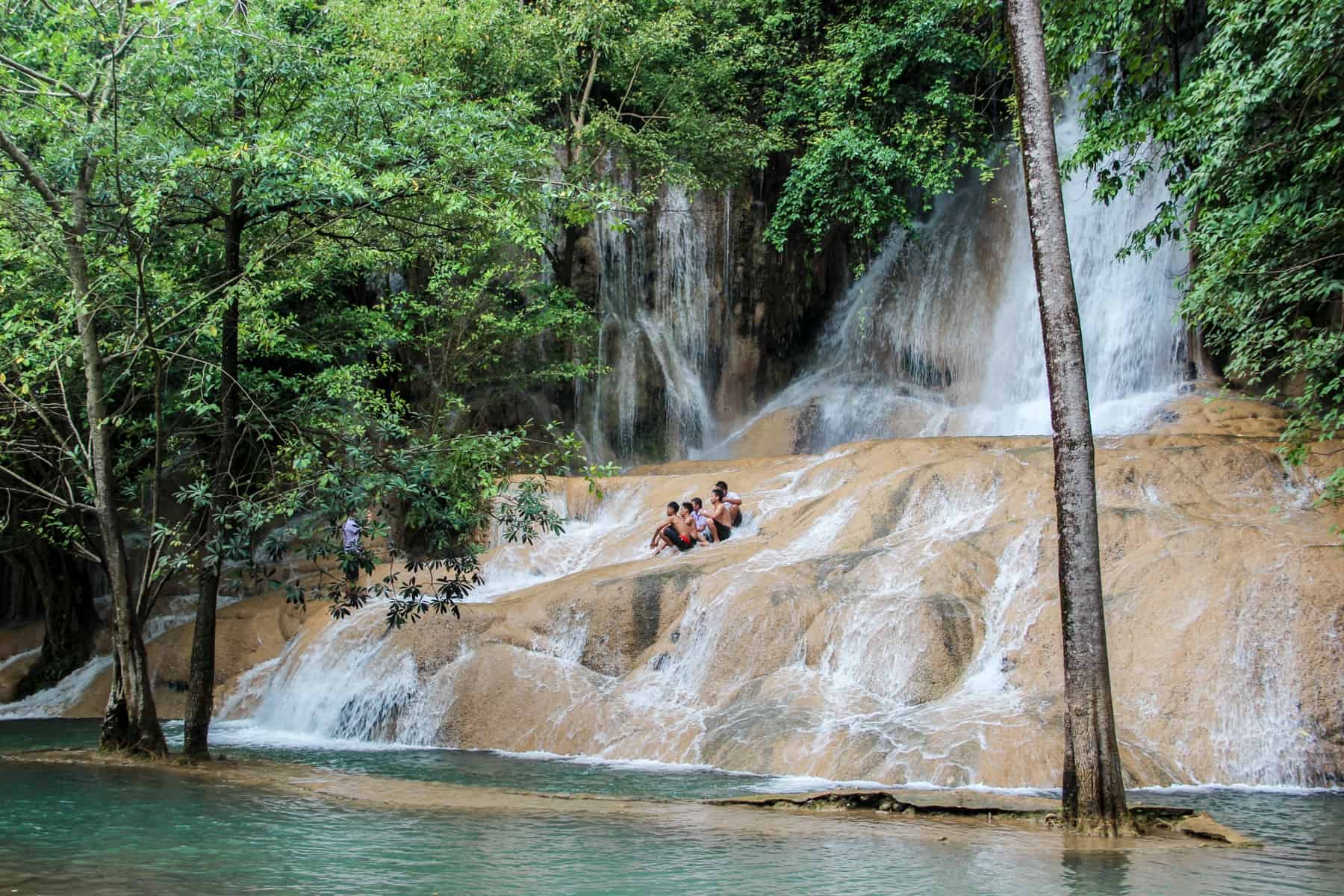 A group of young Thai men sit on a golden limestone rock mound in front of a large waterfall that flows from the forest above