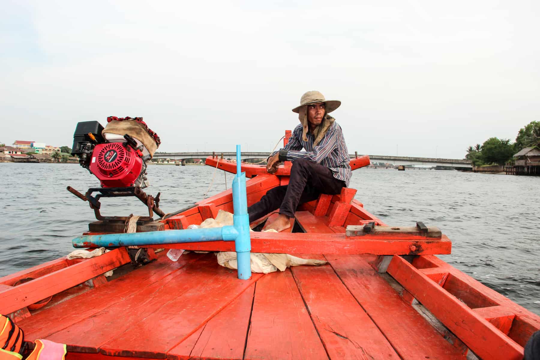 A man in a beige hat steers a red painted wooden boat on the wide river in Kampot in Cambodia. 