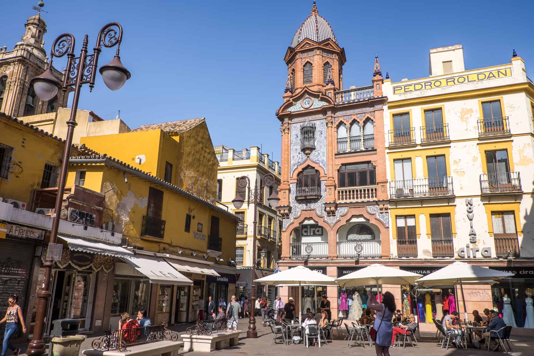 People in a public square in Seville Spain filled with buildings painted bright yellow and vivid salmon pink, vivid in the strong sunlight.