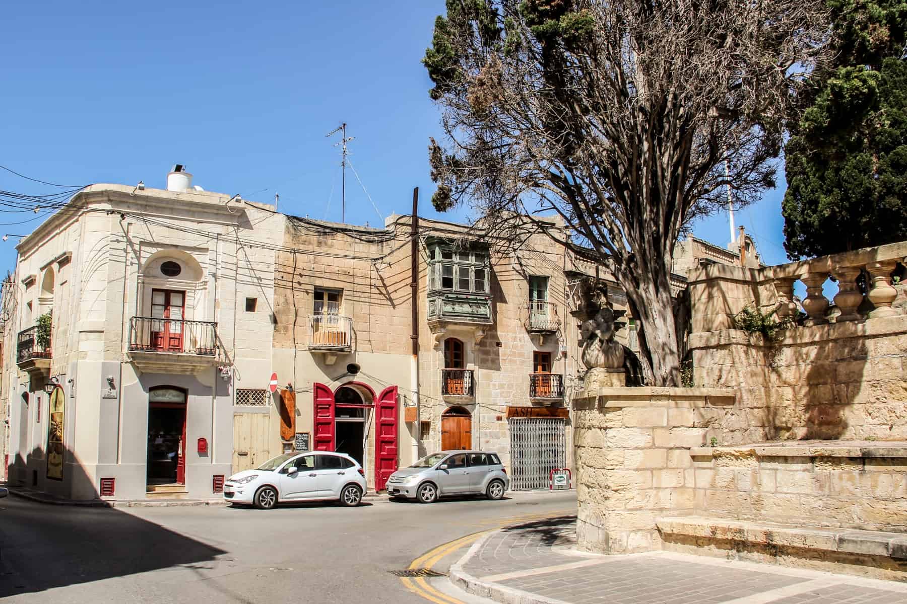 White and silver cars outside a caramel building in Malta, with a red shuttered door and six balconies. 