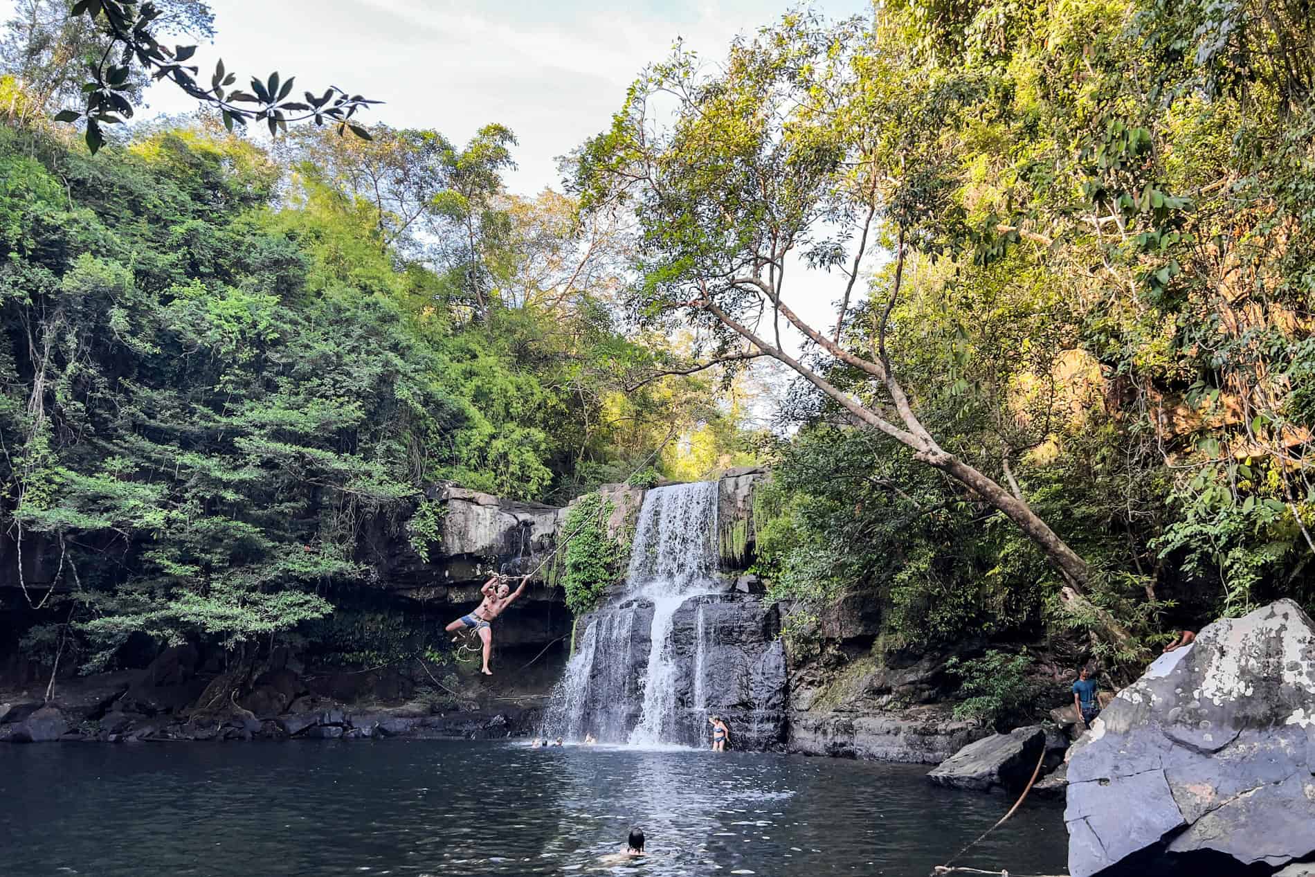 A man swings on a rope, over a body of water in front of a two-tier forest waterfall. 