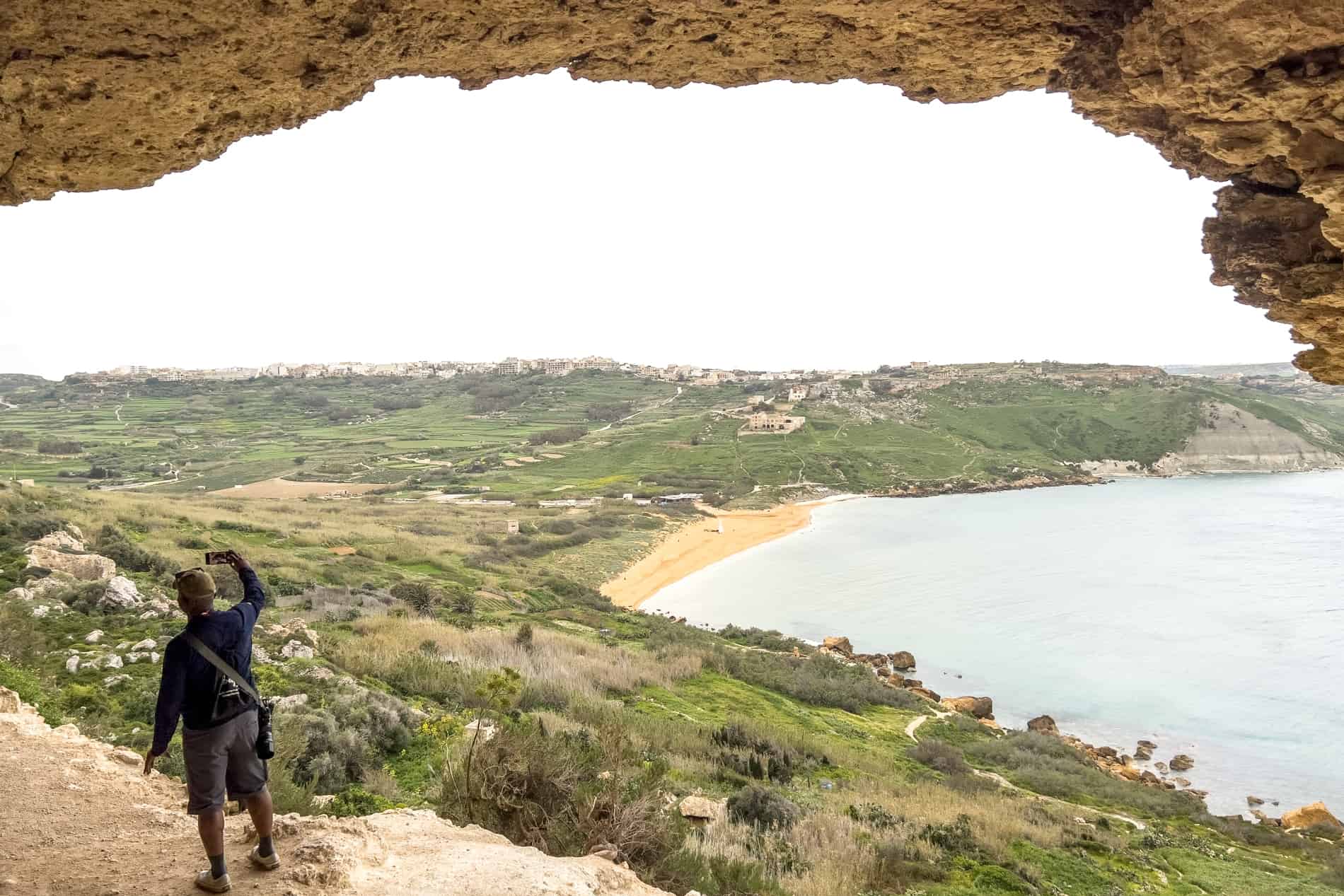 A man take a selfie in a cave in Gozo Island Malta that opens out over a yellow sand coastline backed by forest. 