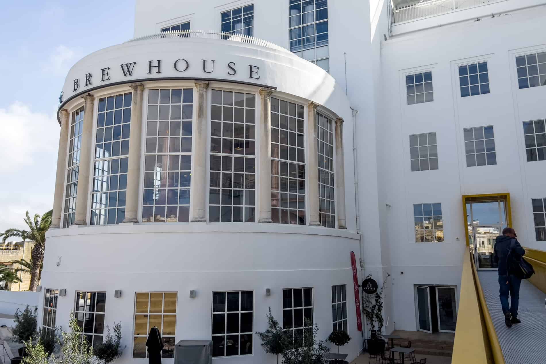 A white building with a yellow ramp and a rounded glass window facade and the words 'The Brewhouse' above it. 