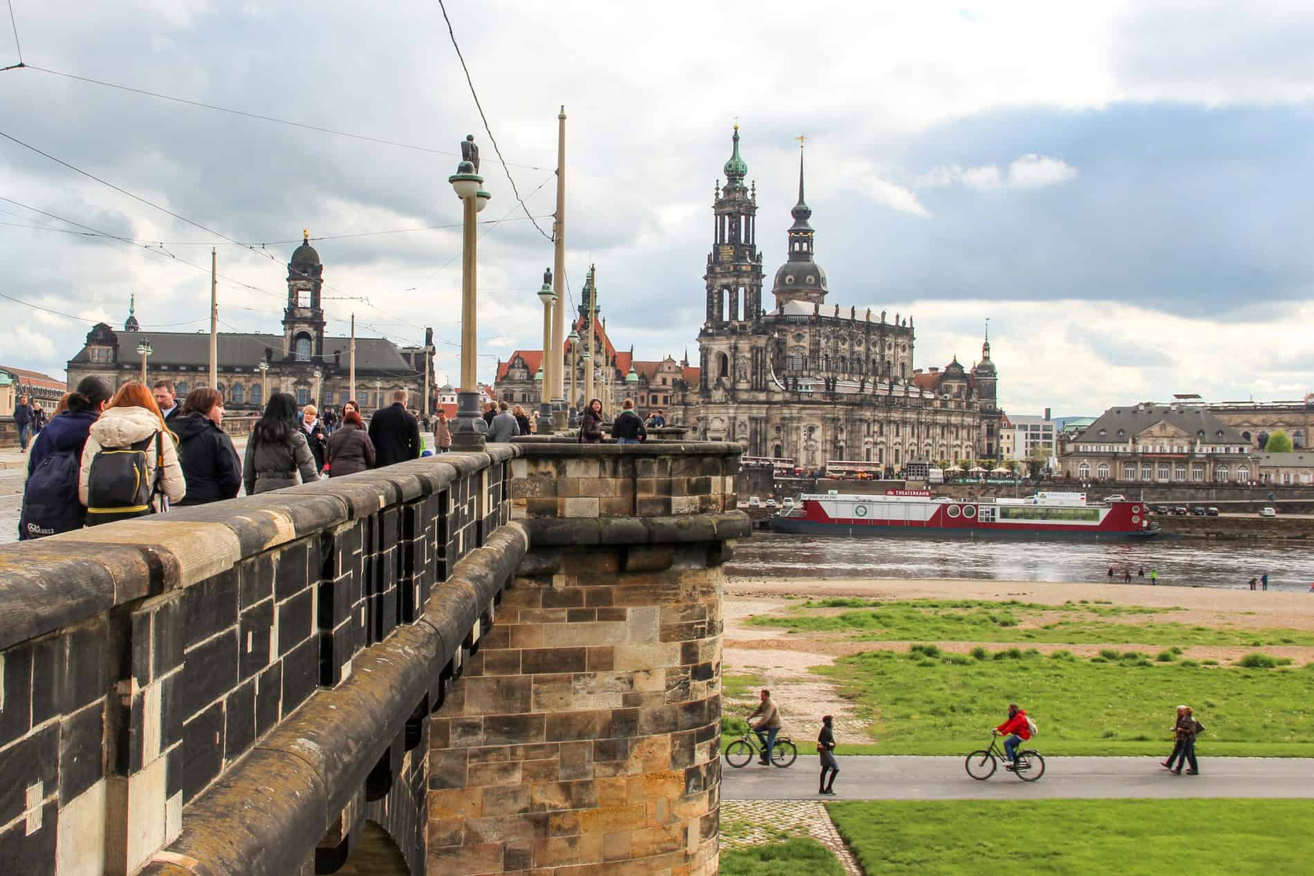 People walking over an old stone bridge towards an opulent spired cathedral complex. Below people walk and ride bikes along the green riverbank. 