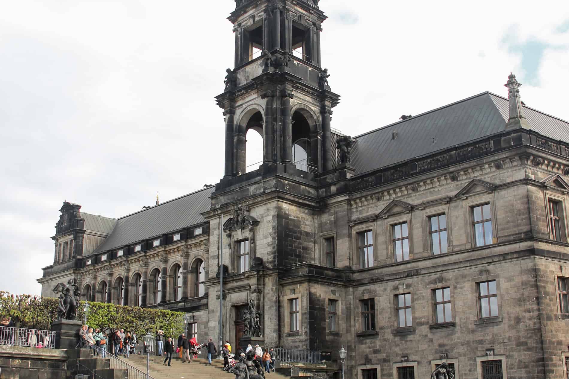 People walkin up the stairs to a rectangular Renaissance building with blackened beige stone and a tall spire. 