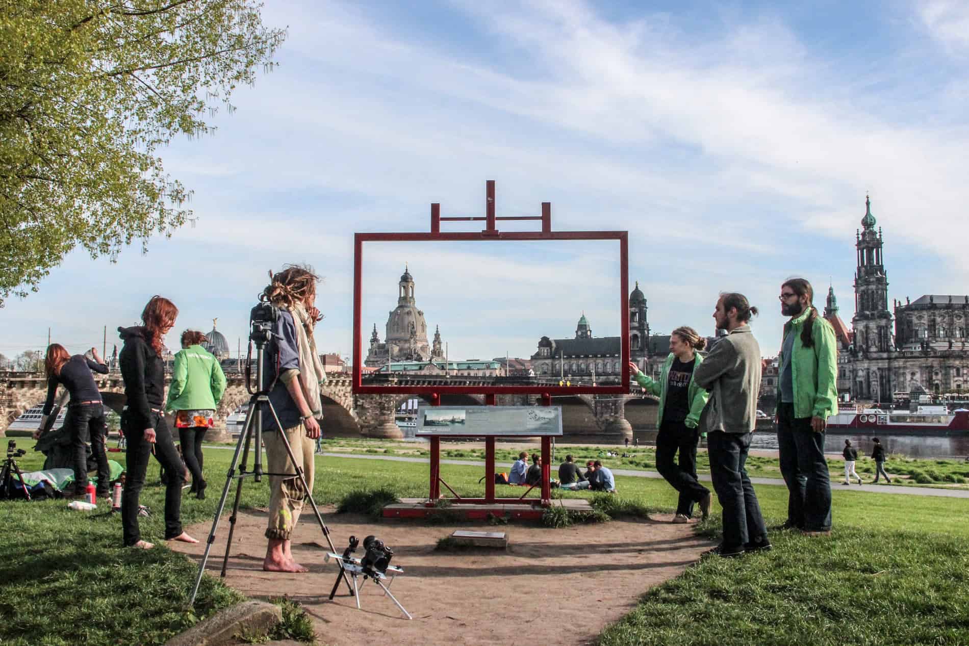 People standing around a giant red picture frame that looks towards (and frames) the old town spires and ornate buildings of Dresden city. 