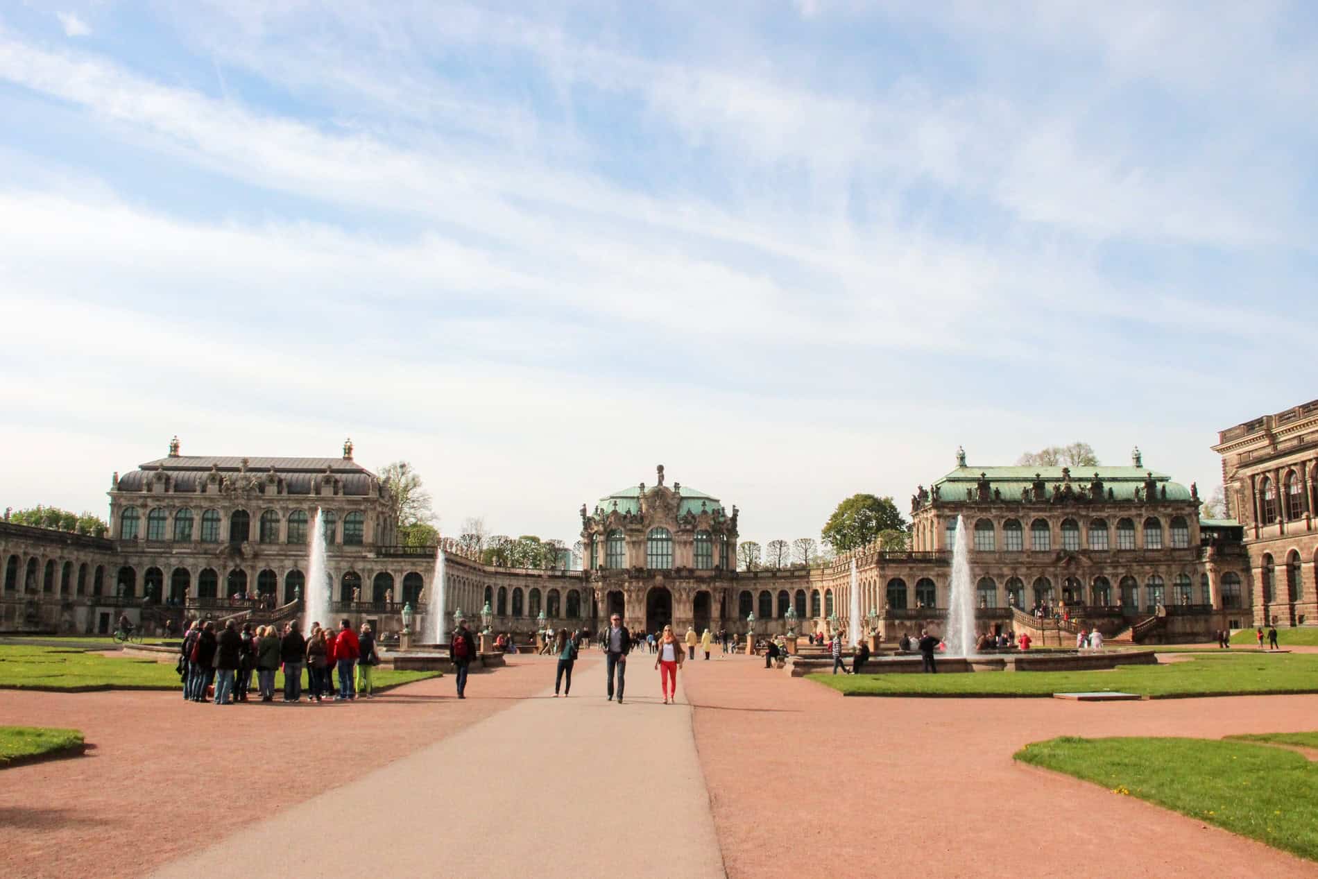 People walking through a large ochre stone and manicured green court yard in front of a long, opulent palace complex. 