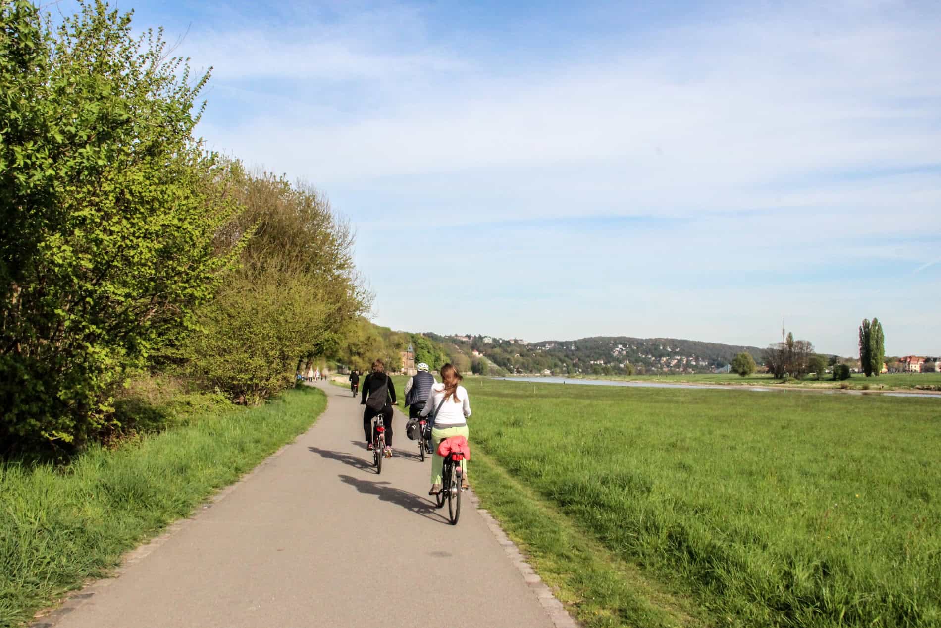 Three people riding bikes on a long path next to a green riverbank. 