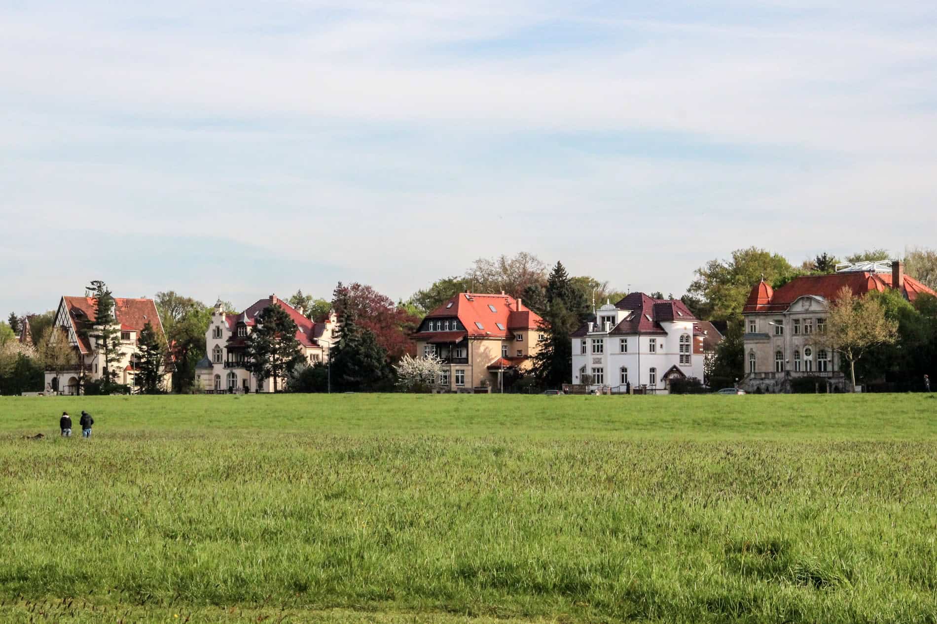 Two people walk in a green field towards a row of five beautiful villa buildings with red-orange roofs. 