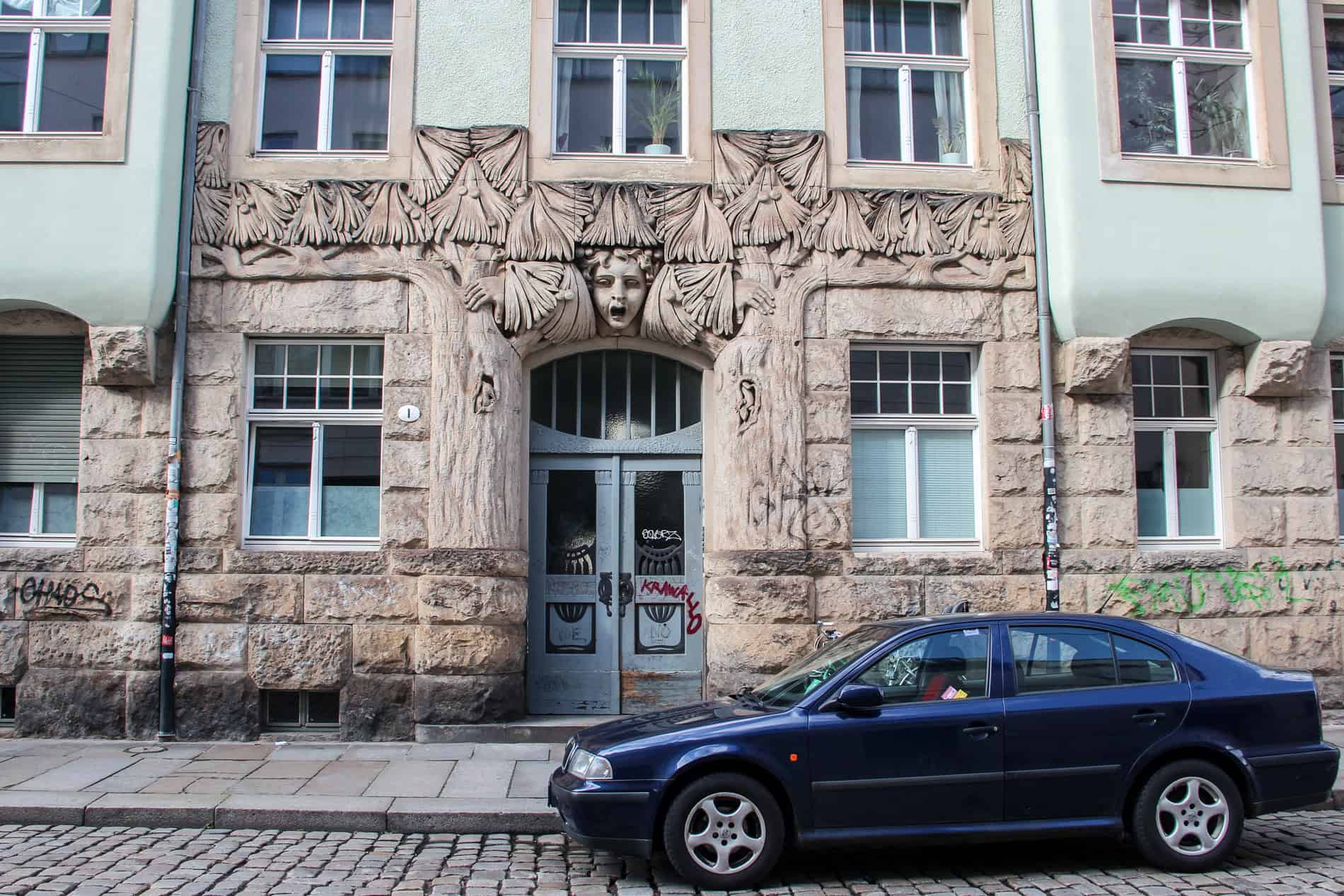 A black car parked outside a building with a stone carved Art Nouveau face design above and around the door. 