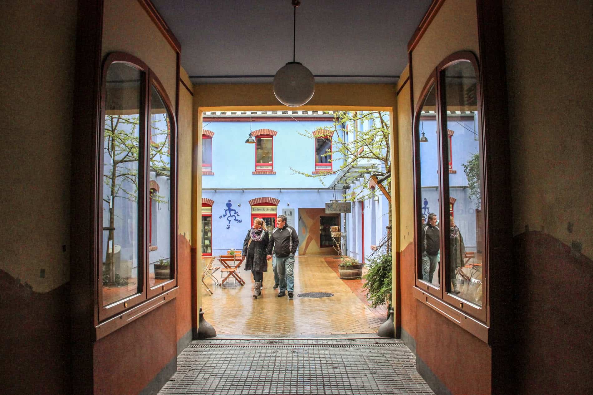 View from a covered passage into a courtyard where a couple walk in front o a blue and purple house. 