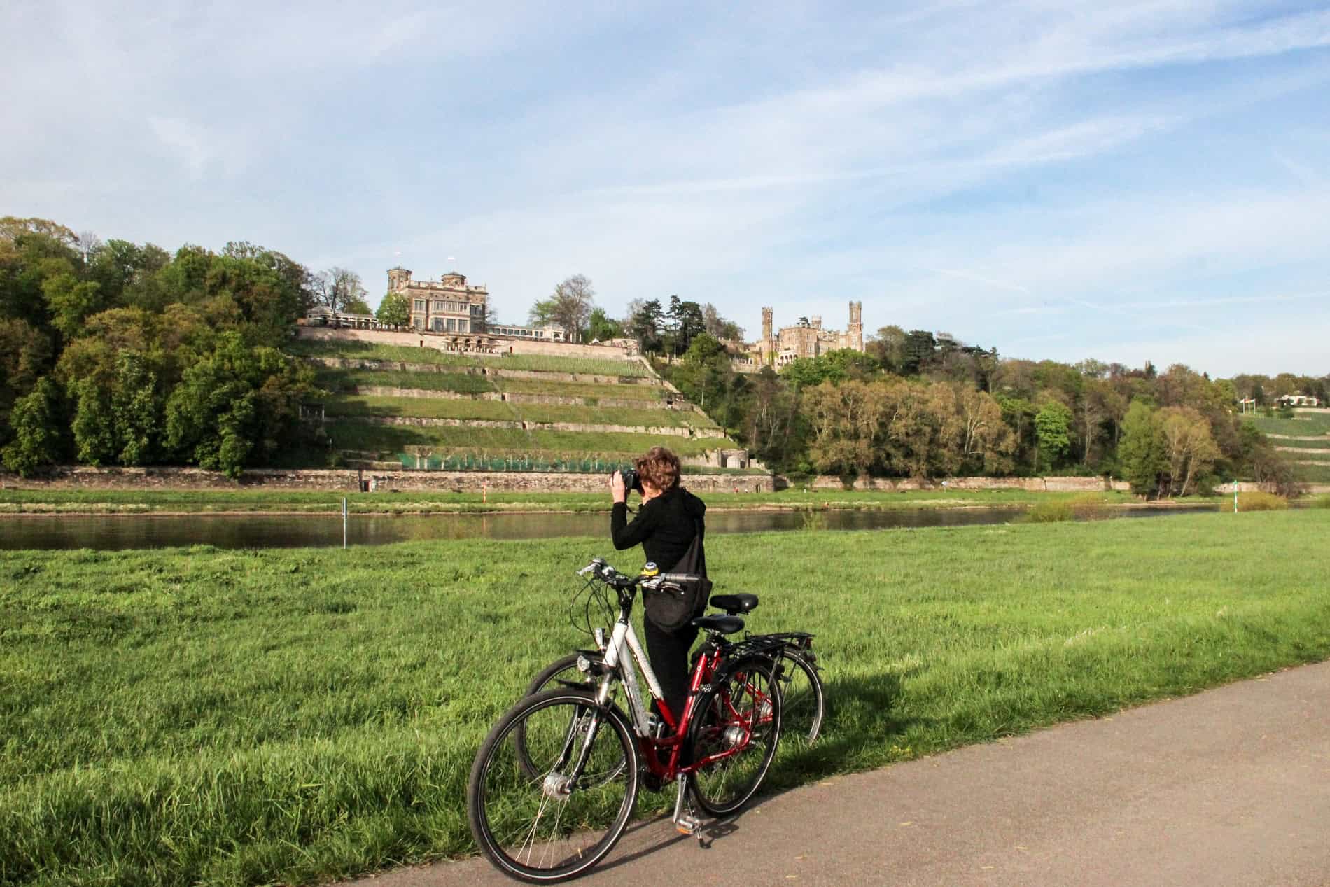 A woman standing next to a bike and taking a picture of a terraced, hilltop castle. 