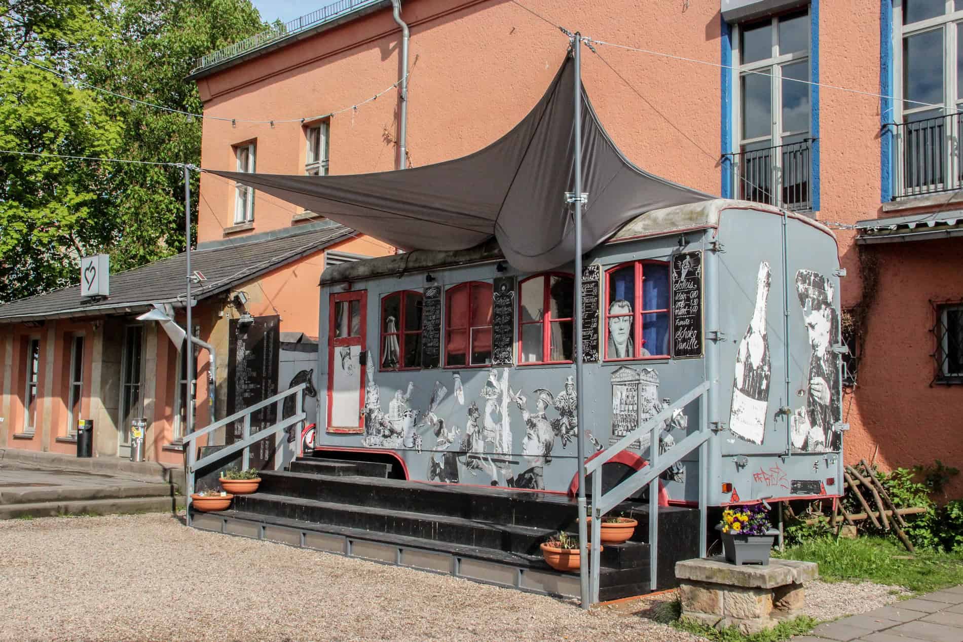 A grey trailer covered with sketches of people, building and wine bottles, outside an orange building. 