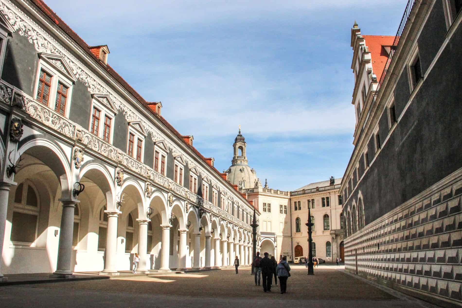 A small group of people walking through a long courtyard, with a long white arched building to the left and a black and white stone building to the right. 