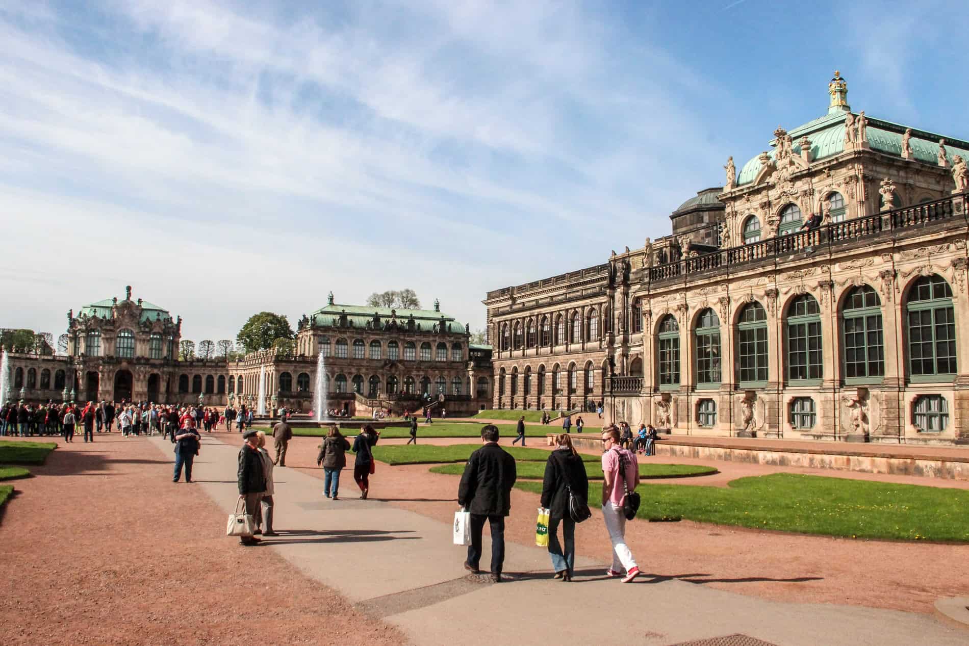 People walking past the decorated stone and mint coloured roof palace complex of Zwinger in Dresden city. 