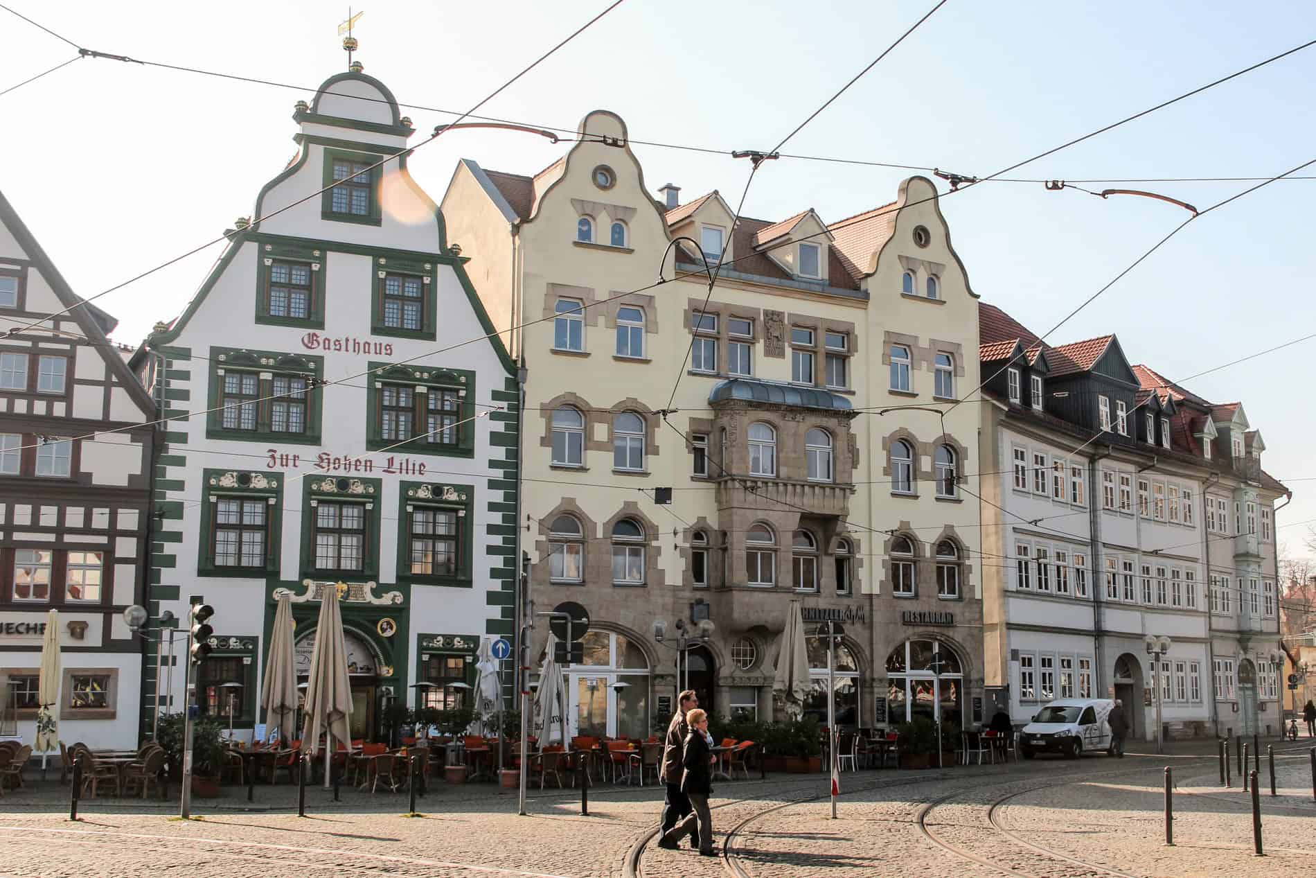 A couple walking on a cobblestoned street in front of a row of thin, muted coloured Renaissance buildings in Erfurt, Germany. 