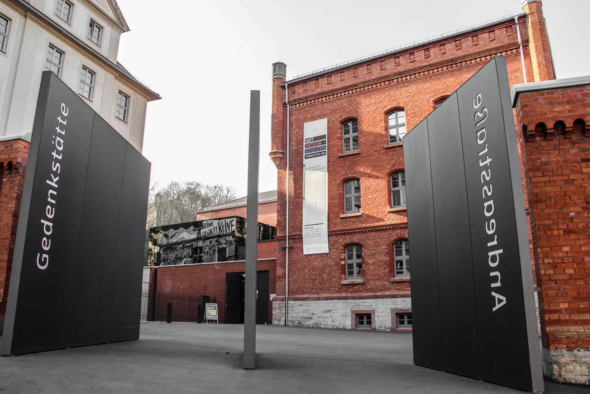 Two thick black doors with the words 'Gedenkstätte' and 'Andreastrasse' open towards an orange brick building – the Erfurt Memorial and Educational Site and former Stasi building. 