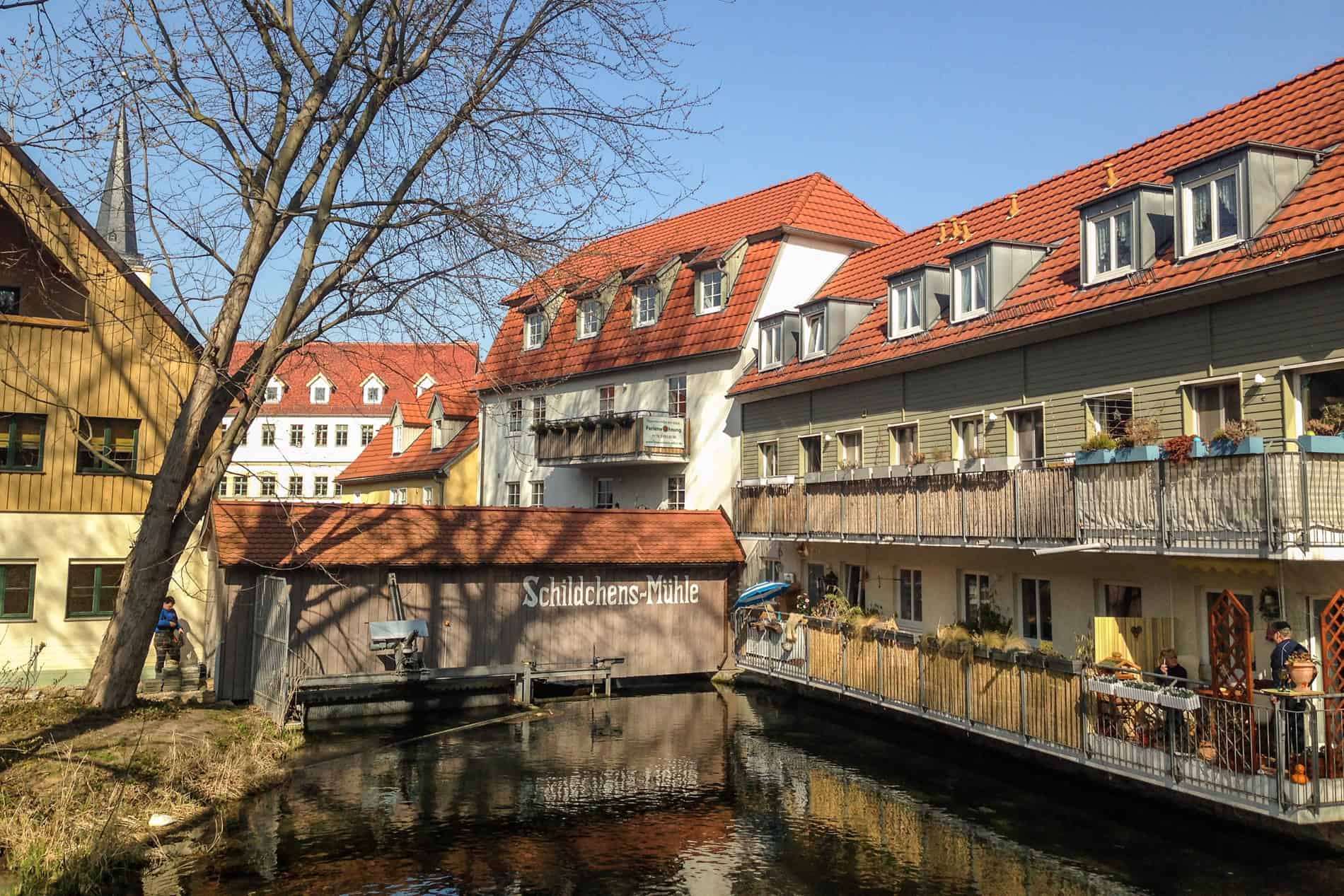 Two people on a orange roofed, green waterside balcony house, connected to a building with the words 'Schildchens-Mühle'. 