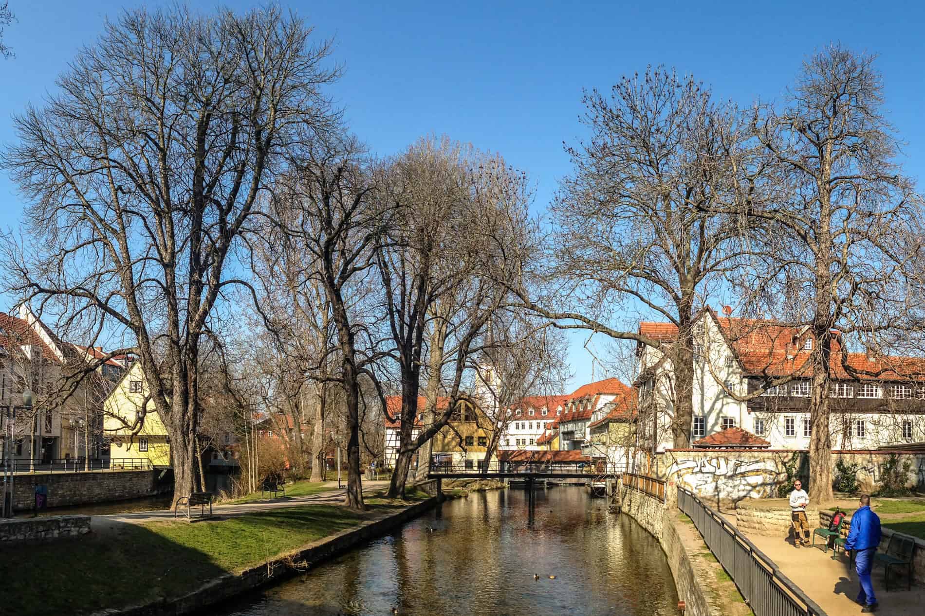 A still and shallow river in front of an old town with orange-roofed buildings. 