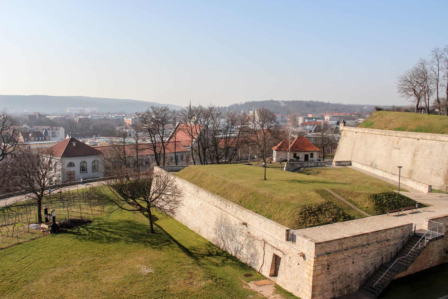 Elevated view over Erfurt city and surrounding hills from the beige stone walls and green grounds of the Petersberg fortress. 